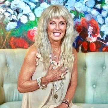Melanie MataJi with long hair, wearing layered jewelry, sits on a couch with a colorful floral painting in the background.