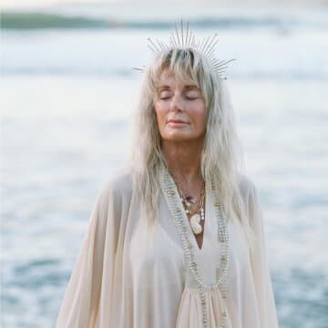 Melanie MataJi with closed eyes and a serene expression, wearing a headdress and flowing garment, stands by the water.