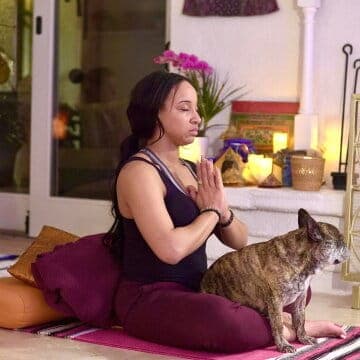 Person meditating indoors with hands in prayer position, accompanied by a small dog. Cozy setup with candles and cushions.