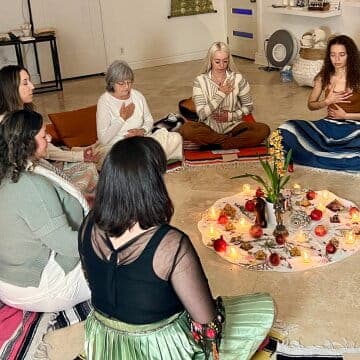 A group of people seated in a circle meditates around a candlelit altar with flowers at The MataJi Sanctuary.